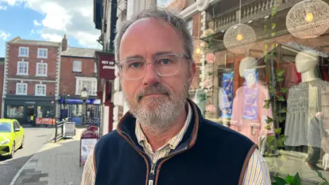 Ellen Knight/BBC Simon Lyon is looking into the camera - he is wearing a yellow and blue striped shirt with a dark blue gilet. He is wearing clear-framed glasses, and has short grey hair and a short beard. He is pictured outside the shop - through the front window you can see two mannequins wearing checked blouses. To the left of the photograph you can see part of the high street. The sky is blue with some white clouds. 