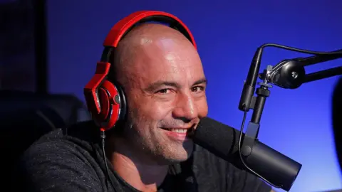 Getty Images Joe Rogan, wearing orange headphones over his ears, smiles into a microphone during a recording of 