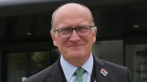 A man wearing a suit and tie smiling at the camera. He is wearing a LGBTQIA+ badge. He is balding and is wearing dark framed glasses, a dark suit, light blue shirt and green tie. 