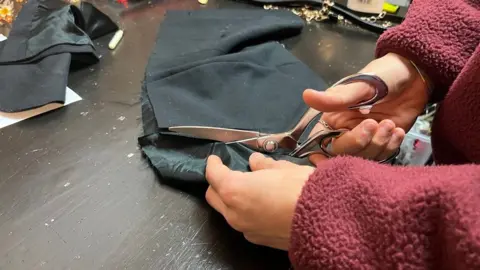 Tom Jackson/BBC Diaconu is cutting a piece of grey fabric on a dark wooden table. He is wearing a red jumper.