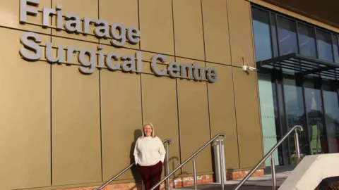 University Hospitals Tees Danielle Gash is a woman with shoulder length blonde hair with a white jumper and dark trousers. She is stood on some steps to a building with a sign that reads Friarage Surgical Hub. It has brown cladding and a few windows.