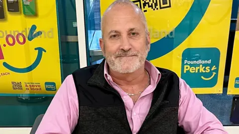 Simon is looking into the camera and has a white beard and bald head. He is wearing a pink shirt with the top buttons undone and a gold neck chain is visible. He is wearing a black sleeveless top over his shirt. Behind him are yellow posters in a shop window