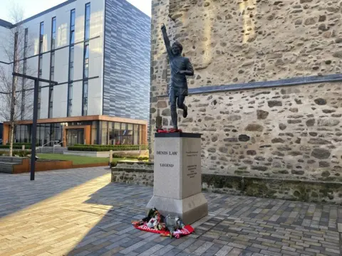 A statue of Denis Law wearing a football strip with his arm raised straight in the air. Football scarves and flowers are left at the base of the statue. It's a sunny day.