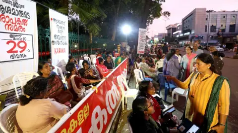 Vivek R Nair/BBC A glimpse of the protest site in front of the Kerala Secretariat where protesters have camped out on white plastic chairs with placards and banners.