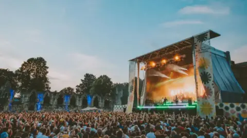 Picture of a previous gig held at Museum Gardens. The stage is seen to the right of the picture, while large crowds are in front. The remnants of a church can be seen in the background.