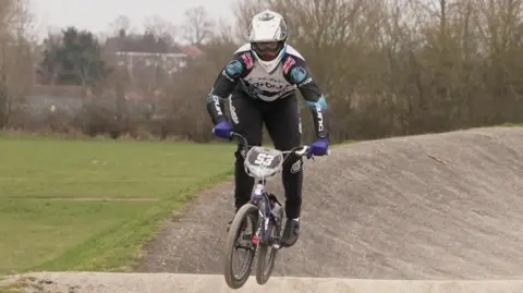 BBC/Shaun Whitmore Jared Hill rides his BMX bike over a jump on his home circuit in Norwich.  He is wearing a white, black and sky blue racing suit and white helmet.