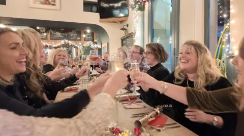 BBC A group of people sit around a long dining table in a bar. There are Christmas crackers on the table and Christmas decorations in the background. The group are holding their wine glasses up in a 'cheers' action. 