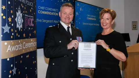 Mark Cooper and Alex Spargo are standing in front of a screen holding a certification and smiling at the camera.