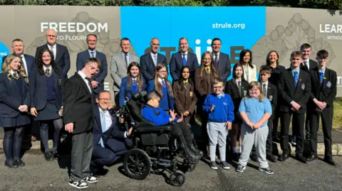 Ten in the back row, eight men in suits including Paul Givan and then two women in blazers.
Fifteen in the front row: left to right is two girls in navy school uniform, one boy in black and grey uniform, a man in a suit kneeling down with his hand on the wheelchair of a boy in a blue uniform slightly in front of two girls in blue uniform, two girls in brown uniform, one boy in blue uniform beside a girl in blue and a boy and girl in navy uniforms.
Behind the two rows is a blue and cream sign that has the world 'strule' on it.