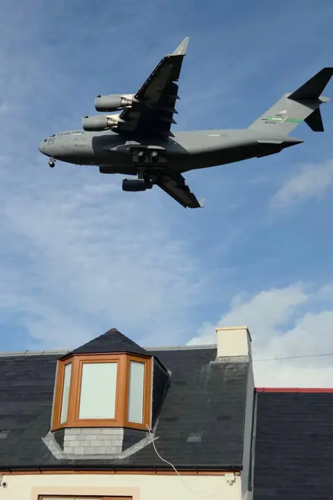 Stu Smith  A large grey US Air Force military jet aircraft against a blue sky above a house with bay dormer window in the roof.