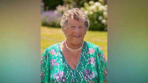 Guernsey Swimming Club Jo Norman is wearing a bright green dress with pink flowers and white spots. and a pearl necklace. There is a garden with purple flowers behind her.