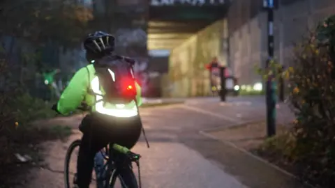 LCC A cyclist riding away from the camera. It is dusk and the bike has lights on. An underpass is visible ahead.