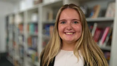 Liv is beaming at the camera. She is standing in the library at MK College. She has long blonde hair and is wearing a white top under a black jacket.