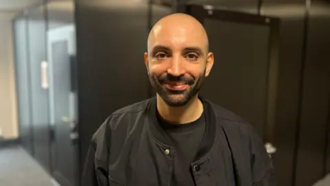 A bald man with a short dark beard smiles at the camera wearing a black jacket over a black T-shirt. He is standing in a corridor with dark glass panels behind him. The background is slightly blurred with warm overhead lighting.
