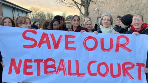 A group of women holding up a big white sign that says 'Save our netball court' in red writing. They are stood outside a leisure centre. 