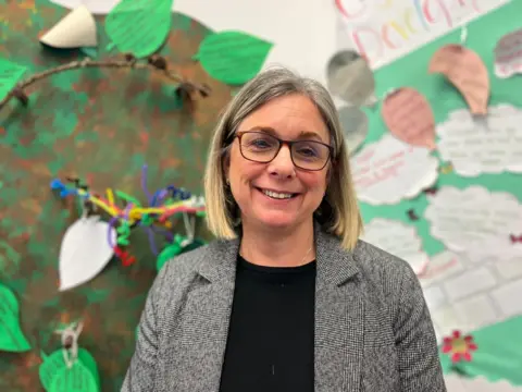 Curriculum manager Anne Clark wearing a pair of glasses, a grey blazer and a black top. She is smiling and standing in front of a colourful nursery artwork wall. 