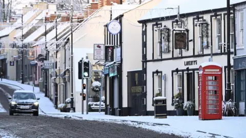 BBC WEATHER WATCHER - Gavan Caldwell A black car travelling down a street covered in snow. There is a pub, red telephone box and traffic lights in shot.