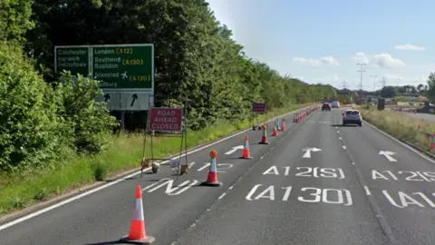 Google The A138, which has three lanes. The left-hand lane is closed and has cones blocking it. There is also a sign that says "road ahead closed" in the carriageway. There is a large road sign showing motorists the different directions they can go in, including to Colchester and London. There are cars in the distance using the middle and right lanes.