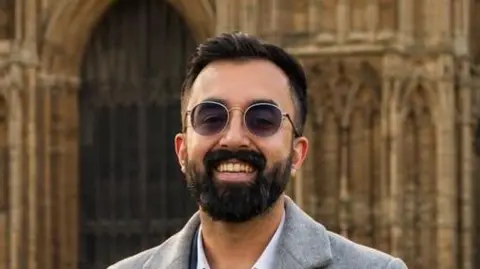 A man smiling straight at the camera while standing outside a cathedral. He is wearing a grey coat over a white shirt and has sunglasses on.