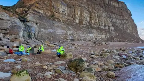 HM Coastguard A distanced view of four coastguards, in yellow hi-vis jackets, tending to several people who are trapped beneath cliffs at a pebbled beach. 