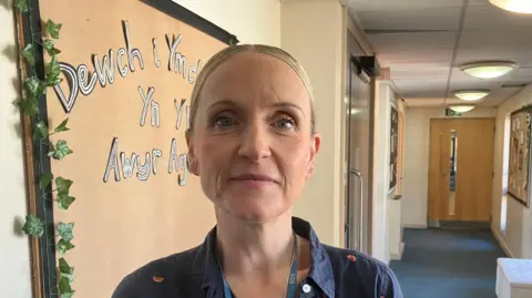 A woman with pulled back blonde hair wearing a blue shirt with a watermelon slice pattern, stands in a school corridor. She is wearing a lanyard with an NHS logo ribbon.