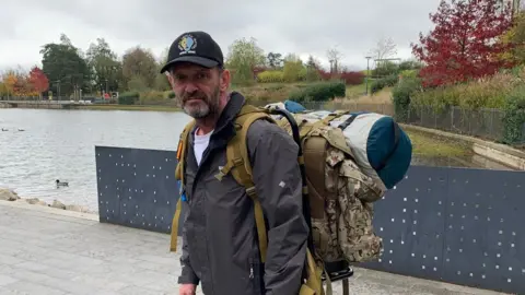 A man with a dark beard in a grey jacket and black cap with a large backpack, standing in front of a small lake with a metal fence