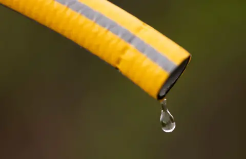 A yellow hose with grey stripes pointing diagonally downwards with a drip of water coming out the end