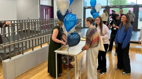 Students pictured in a school hall, collecting their results. Three girls are at a table. There are navy, blue and white ballons.
Behind them is a camera man and some parents.