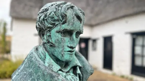 Emma Baugh/BBC Weathered looking greenish statue of John Clare. Its head is turning sideways and looking down, has deep eyes and sideburns. Behind it is a low-lying white painted building with black painted windows. 