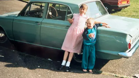 Family handout A young girl and boy stood in front of a blue car smiling. 