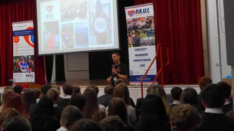 Paul for Brain Recovery A man delivers a talk to pupils inside a school hall, there is a slide projector and several banners on the stage.
