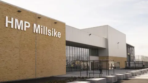 The front of a modern prison with a high brick wall giving way to a modern-looking frontage made up of black glass and grey cladding. A sign in large white letters reads "HMP Millsike".