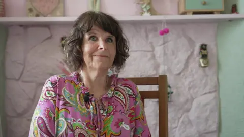 Tiffany is sitting on a wooden chair, smiling at the camera. She is in front of a pale pink and green wall. Her brunette hair is in a short bob and she is wearing a pink top with colourful swirls and shapes on.