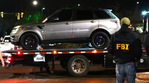 An FBI agent, with his back to the camera stands in front of a vehicle being towed. He's wearing jeans and a black jacket with FBI written on it