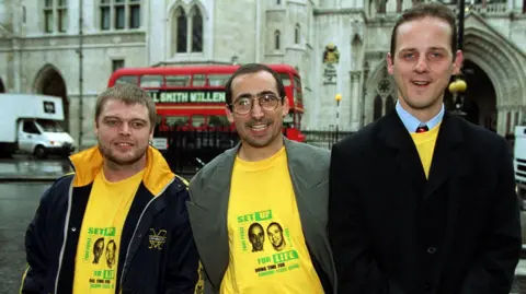 PA Media Three men stand in front of the Royal Courts of Justice in London with a red London bus also behind the,. They are smiliang wearing yellow tshirts