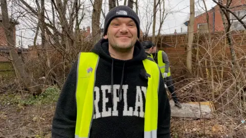 Man in a black hat, black woollen hoodie and bright yellow hi-vis smiling.