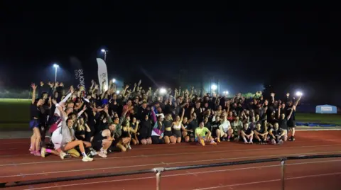 John Fairhall/BBC The Harmonious Wellness Club pose together on their track night event at an athletics circuit. There are around a couple of hundred people huddled in a group in their running gear cheering, with their arms in the air. Many of the attendees look to be in their 20s and 30s. The track is lit by floodlights.