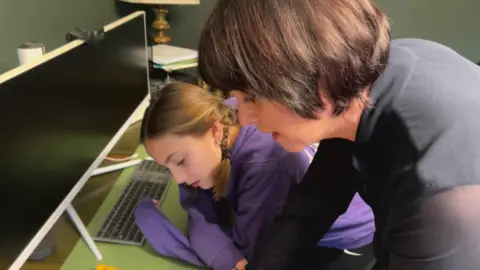 Nathan Turvey/BBC A picture of Ava (left) wearing a purple hoodie and her mum Lucy (right) who has brown hair and a dark top, working at a desk with a computer screen and keyboard.