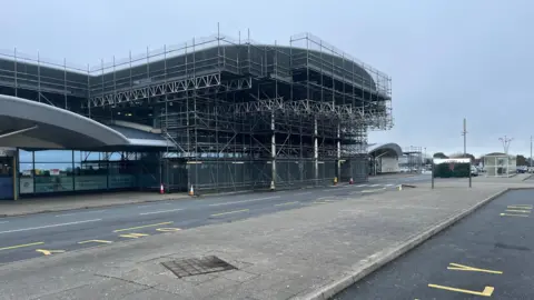 BBC An airport covered in scaffolding with yellow markings on the road in front of it. The sky is grey.