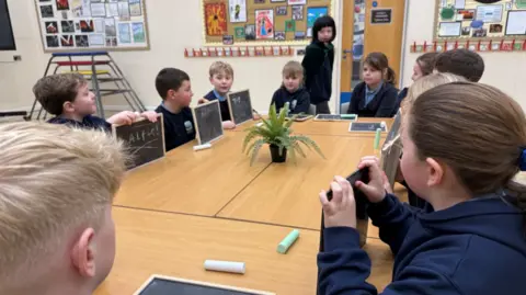 Pupils gathered around a table with chalkboards. With a pupil dressed as Claudia Winkleman, in a black white and green cloak, walking around the table.