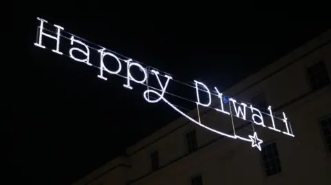 Warwick District Council A neon light hangs between buildings, with the text 'Happy Diwali'.