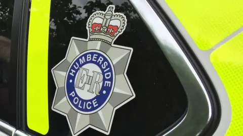 BBC The Humberside Police logo with Humberside Police in a blue circle, ER in the middle and a star and crown around it. Located on the side of a police car