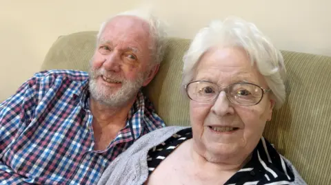 Rebecca Brown Maurice and Velma Brown, sitting on a dark beige sofa at Baedling Manor care home in Bedlington, Northumberland. They are smiling. Maurice has white hair and a grey beard, sideburns and moustache. He is wearing a blue, white and pink checked shirt. Velma is wearing a black dress or top with white stripes, and a beige cardigan. She has glasses and short, white hair.