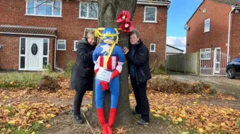 Two women hug a tree on a residential street. They are both wearing black winter coats and black trousers. There is a large super-hero doll tied to the tree.
