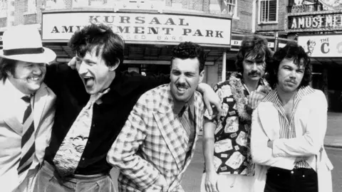 Michael Putland/Getty A black and white image of the Kursaal Flyers outside the Kursaal. They are all wearing funky outfits and smiling while arm in arm. There are five of them.