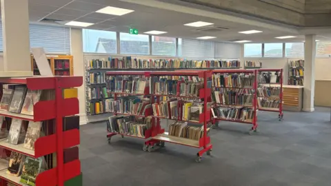 Essex County Council Rows of books on shelves. The shelves are red and the carpet is grey. 
