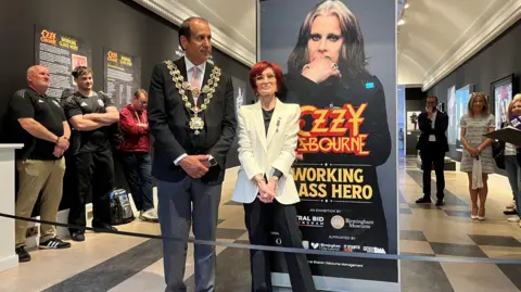 A man and woman stand in an exhibition hall, in front of a poster of a man that says "Ozzy Osbourne, Working Class Hero" on it and a black ribbon. The man is tall and wears grey suit trousers, a black suit jacket, white shirt, pale pink tie and a mayoral chain. The woman has dyed red hair, glasses and is wearing a white suit jacket with a black top underneath and black trousers. There are people standing around the edges of the exhibition hall looking on.