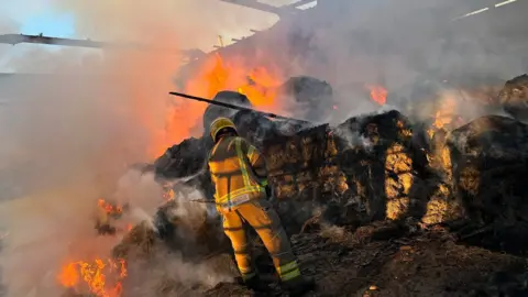 A firefighter pictured from behind in a barn, stood in front of hay which has flames coming out of it and smoke billowing out. The roof of the building has been burnt away.