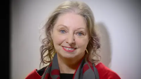 Hilary Mantel in a red top and red/grey scarf, posing and looking at the camera, in 2020.