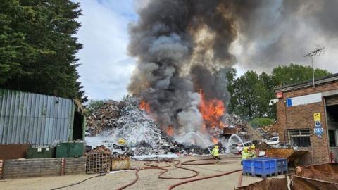 Firefighters crouch in front of a towering pile of scrap metal holding water hoses to douse the flames